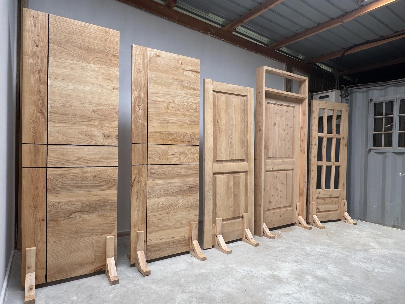 Solid timber doors standing in the TimberStone workshop in Nanyuki, including two tall hardwood entrance doors, a panel door, a door in its frame with a fanlight, and a glazed door