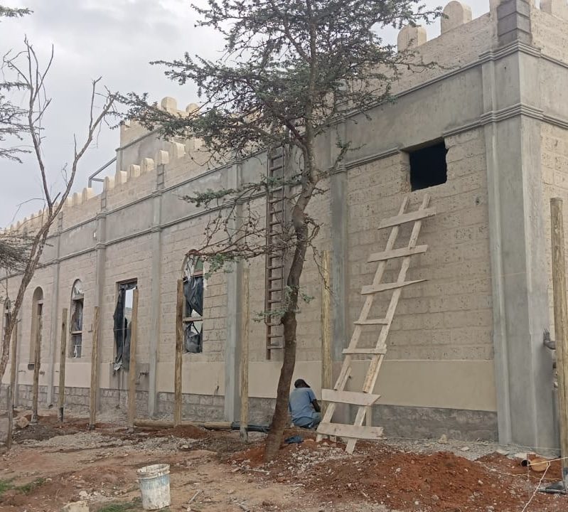 Side wall of the Lolldaiga pool house in October 2026, with window openings, plastered base course and a worker at the foot of the wall.