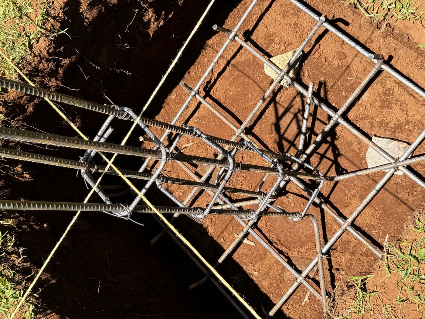 Column cage of vertical bars and bent stirrups sitting on a square mat of footing reinforcement in a red-soil excavation